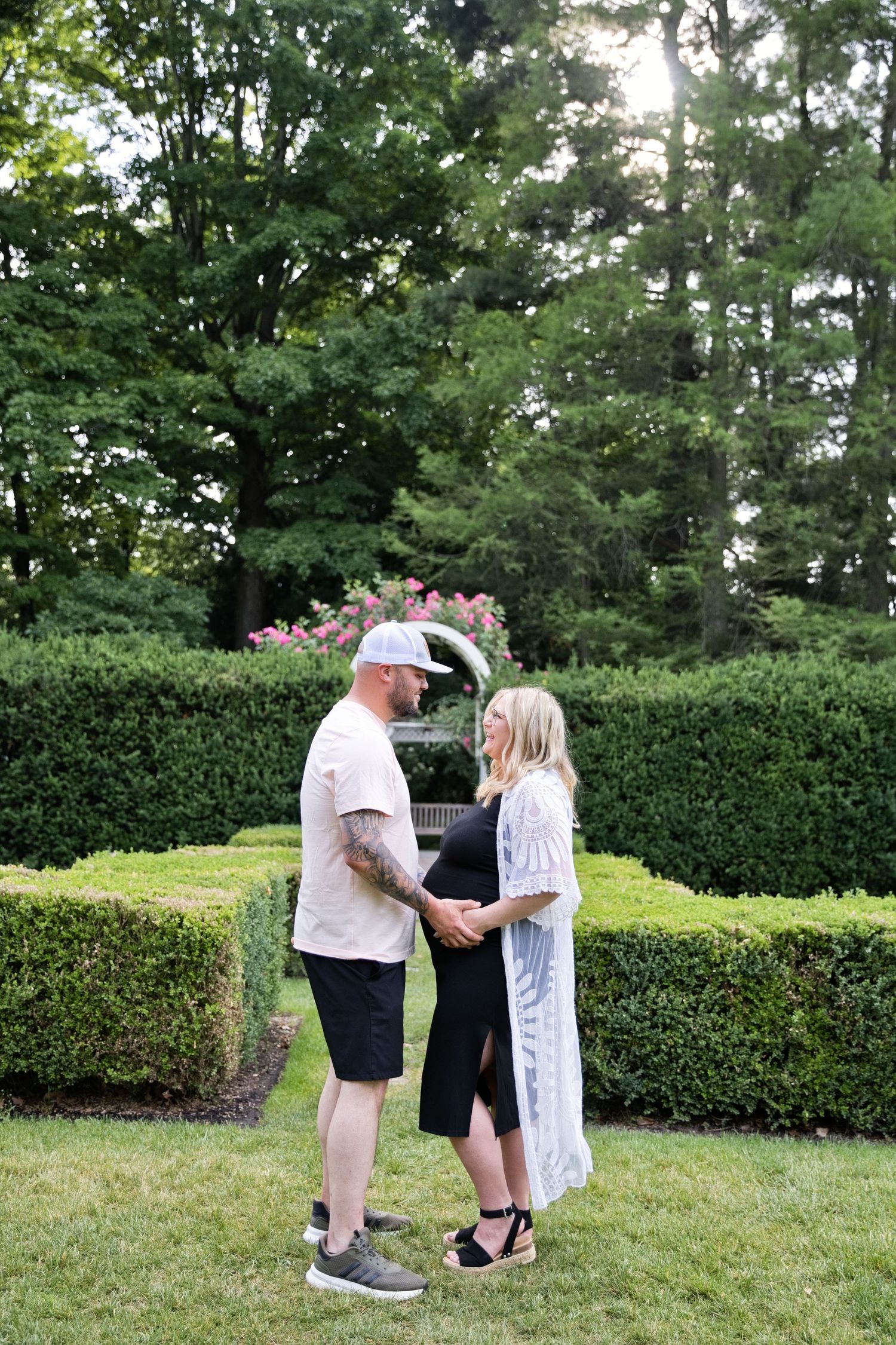 Couple holding hands in a lush garden, pregnant woman in black dress and white cardigan with partner in casual attire.