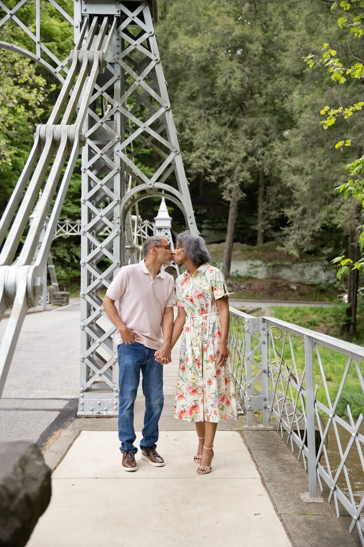 Older couple holding hands and sharing a kiss on a decorative metal bridge surrounded by lush green trees.