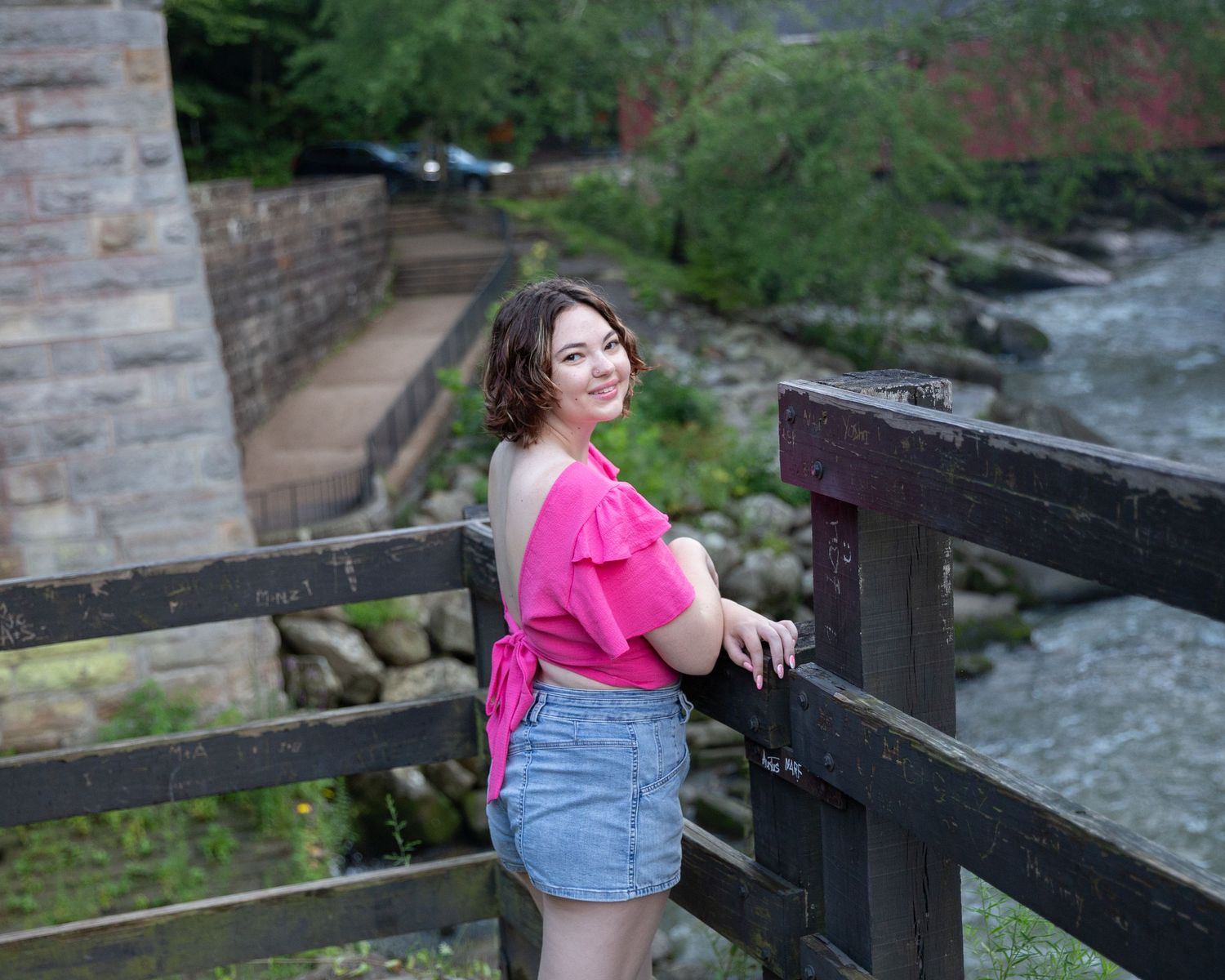 Person in pink shirt and denim sitting near stone wall by flowing water.
