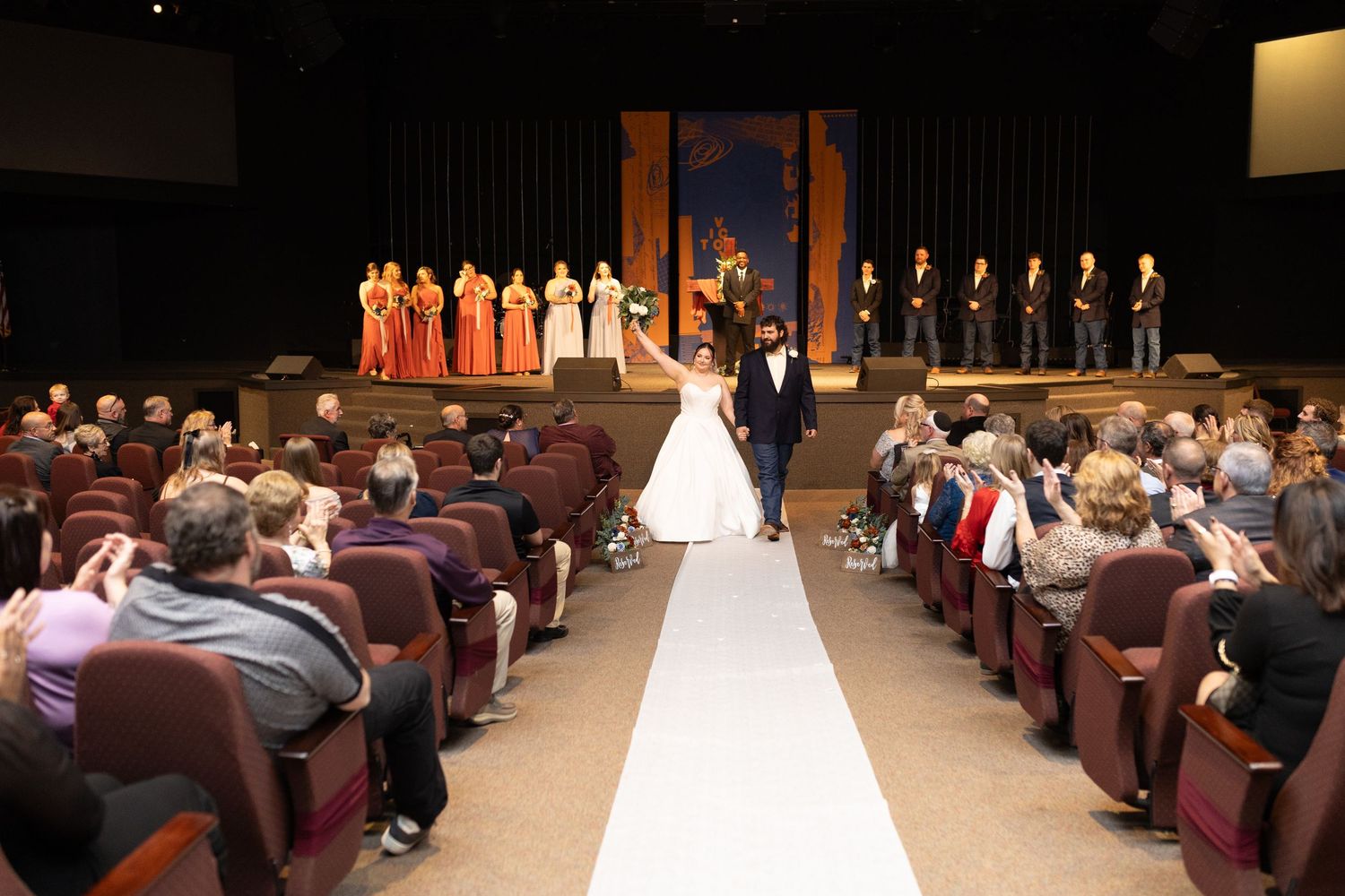 Wedding ceremony in auditorium with white aisle runner and guests seated in theater-style chairs.