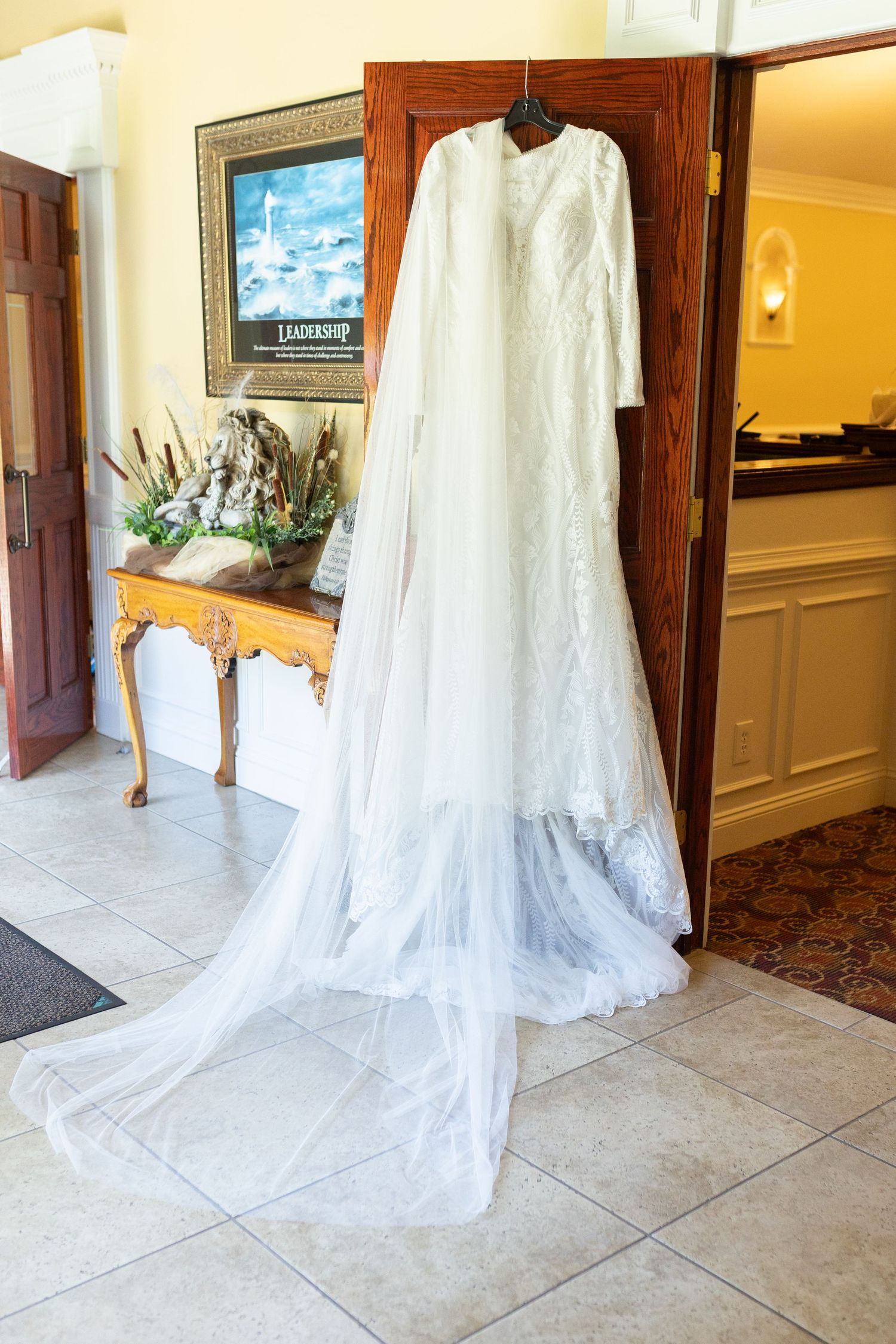 An elegant white lace wedding dress with long sleeves and flowing train hangs on display in an entryway.