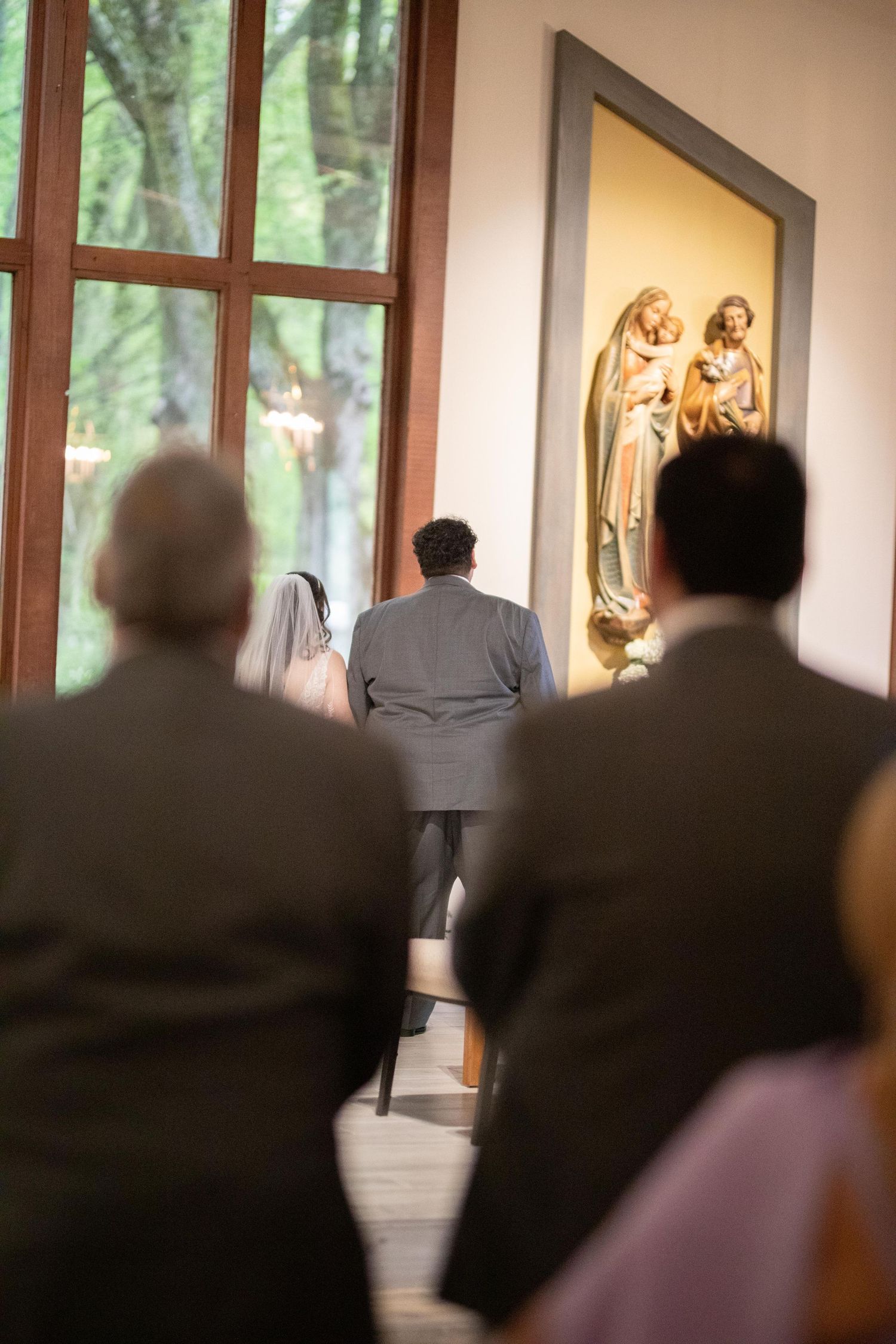 Bride and groom kneeling at altar during Catholic wedding ceremony with religious statues and guests seated behind.