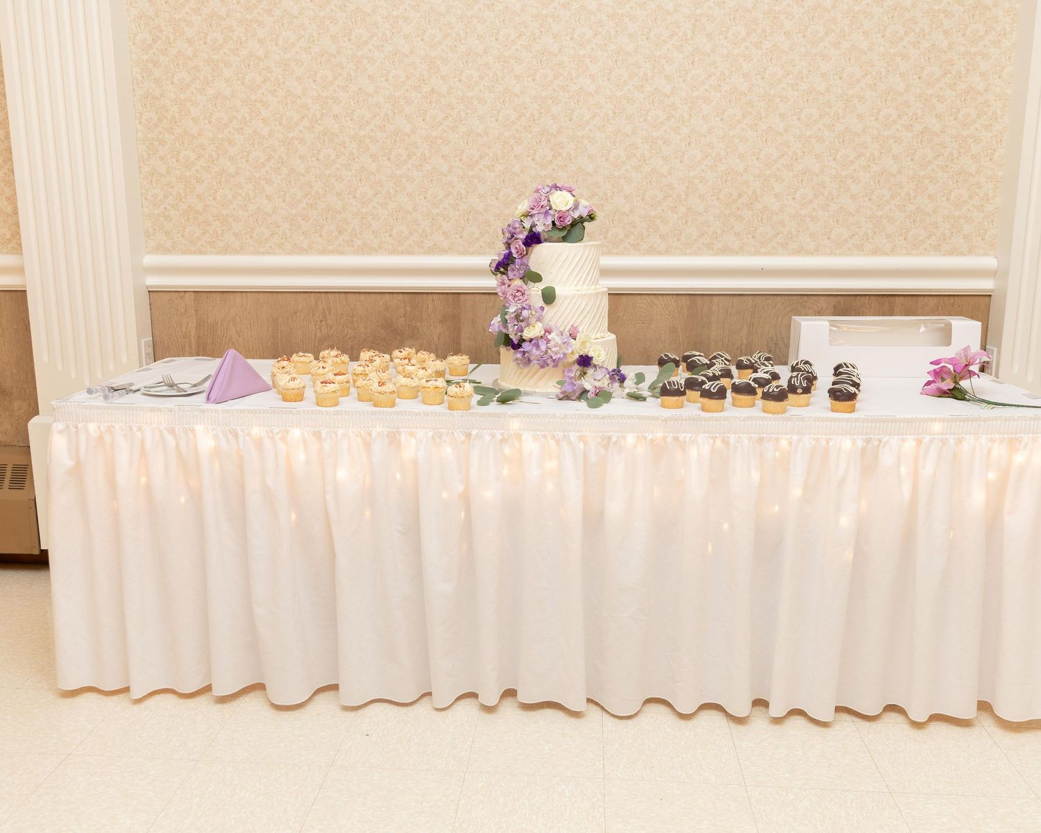 Elegant wedding dessert table with white ruffled skirt, tiered cake with purple flowers, cupcakes, and donuts.