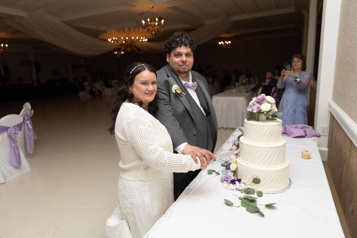 Bride and groom cutting their white tiered wedding cake in an elegant banquet hall decorated with purple accents.