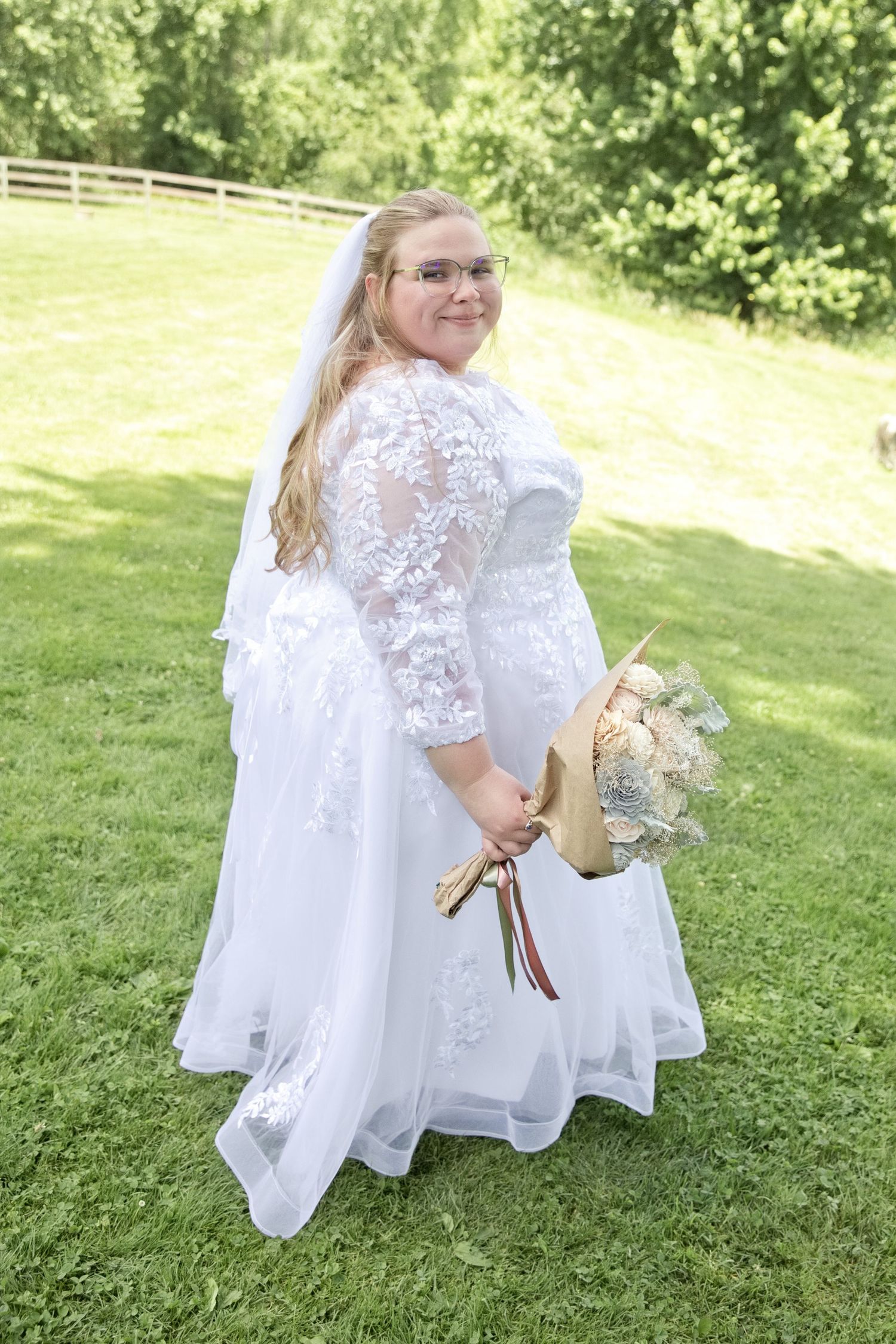 Plus-size bride in white lace wedding gown with veil and bouquet posing outdoors on green grass.