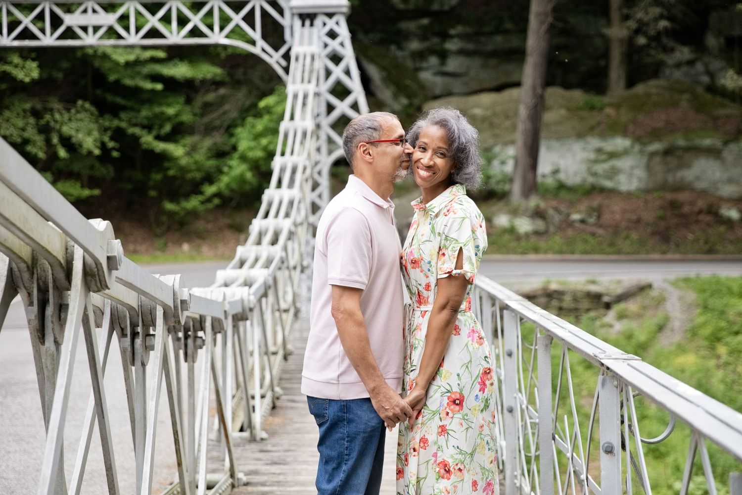 Older couple kissing on a white bridge surrounded by lush green trees and rocky cliffs.
