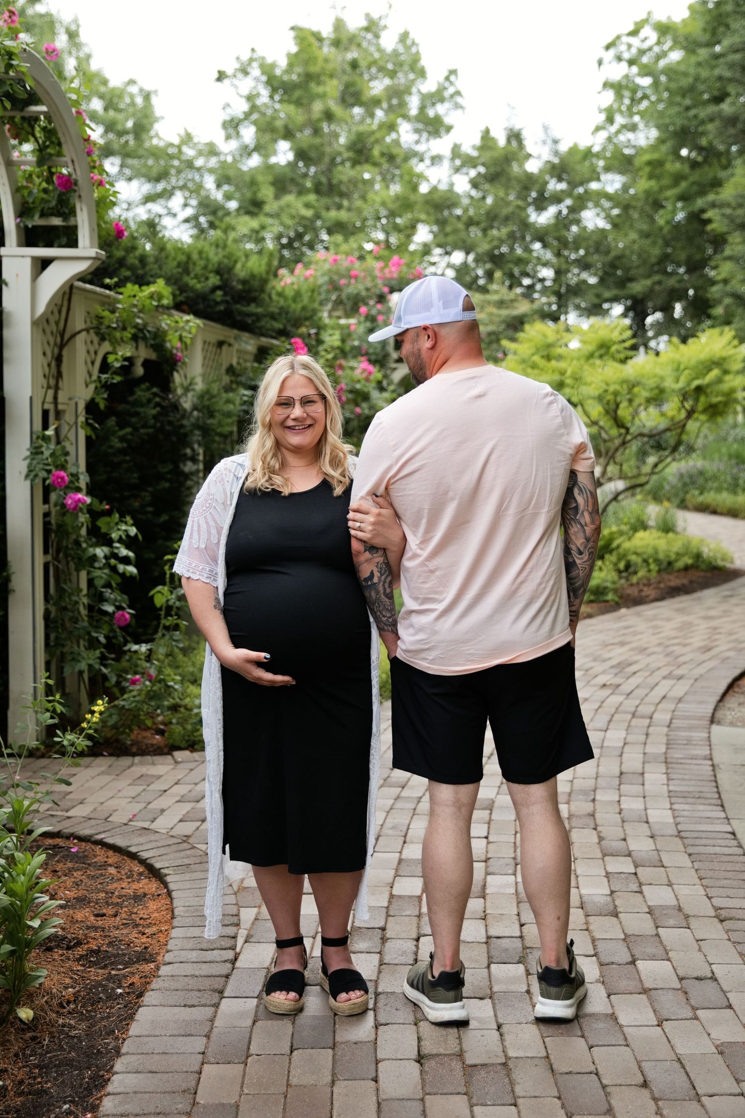 Pregnant woman in black dress with partner on garden path surrounded by lush greenery and roses.