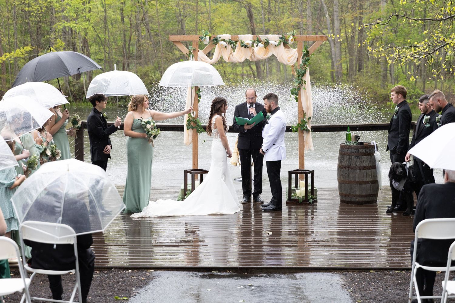 Outdoor rainy wedding ceremony with guests holding umbrellas near a wooden arch by a pond.