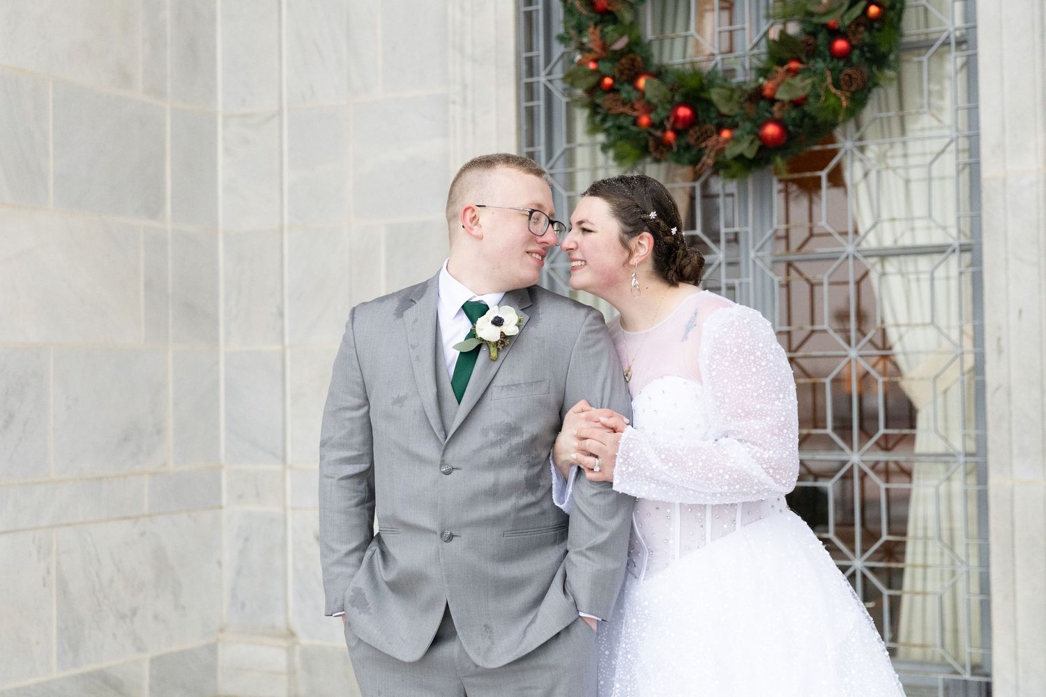 Bride and groom sharing a romantic moment outdoors near a stone wall with Christmas wreath decoration.