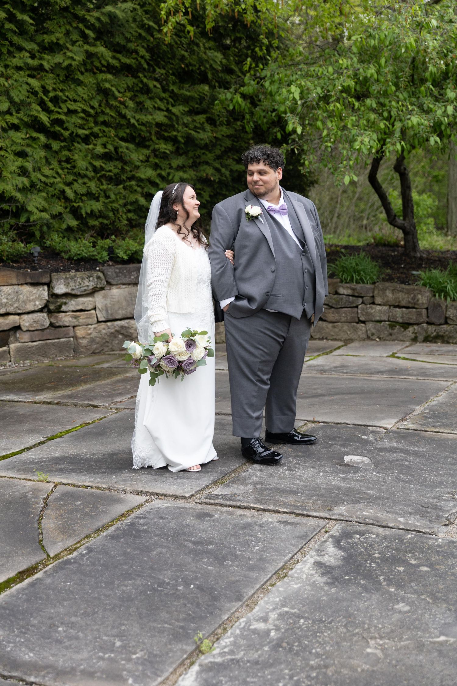 A bride and groom pose together on a stone patio surrounded by lush greenery on their wedding day.