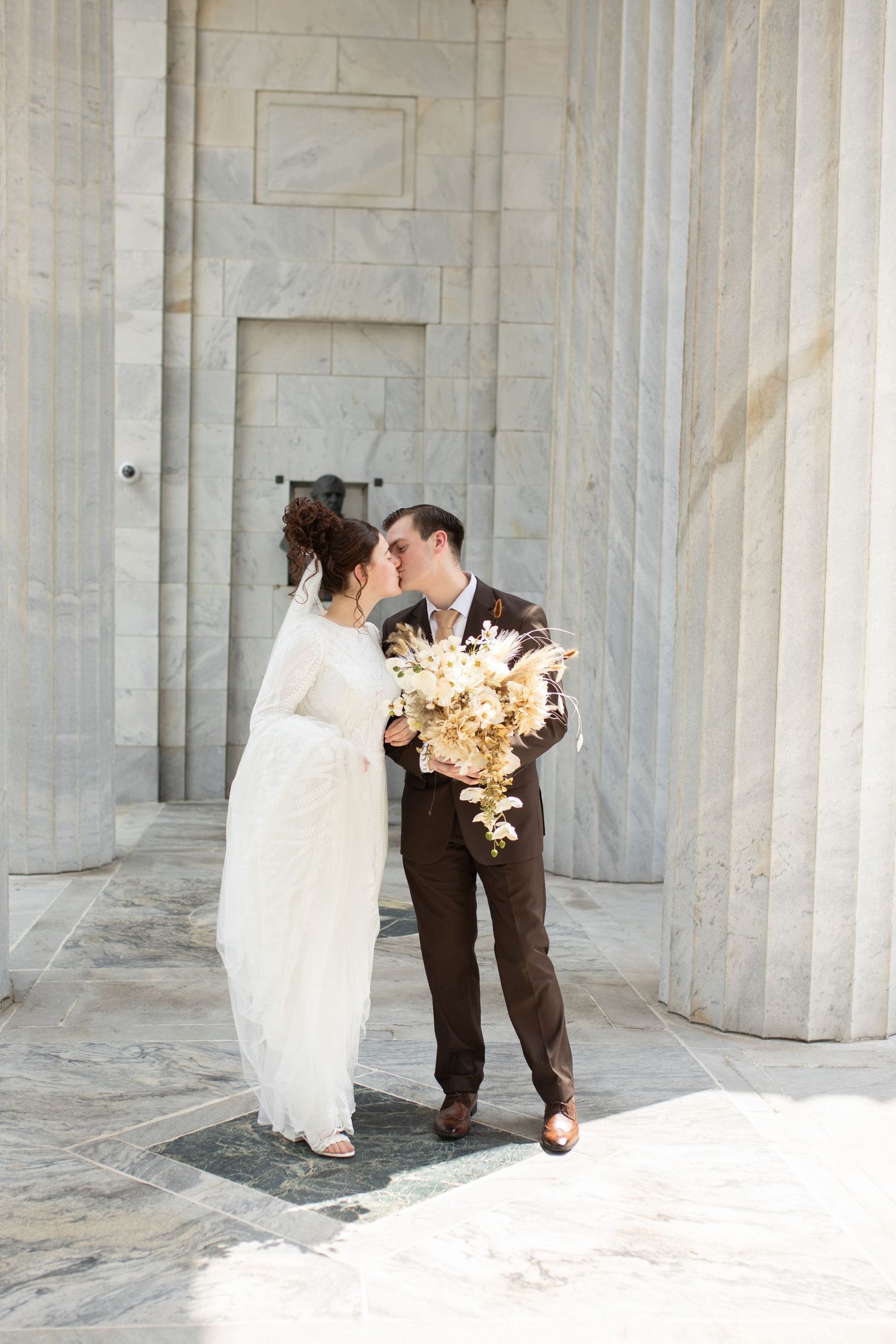 Couple shares intimate moment between marble columns with bride holding neutral floral bouquet.