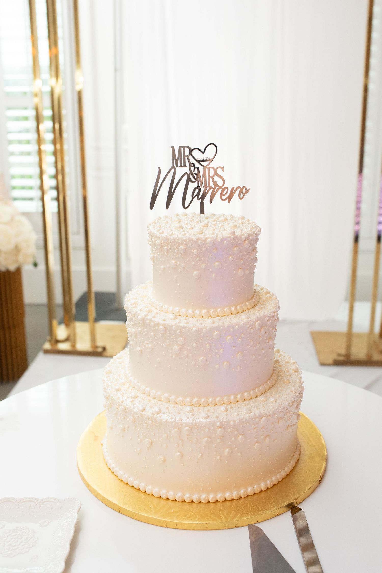 Three-tiered white wedding cake with delicate pearl details and 'Mr & Mrs' topper on gold base.