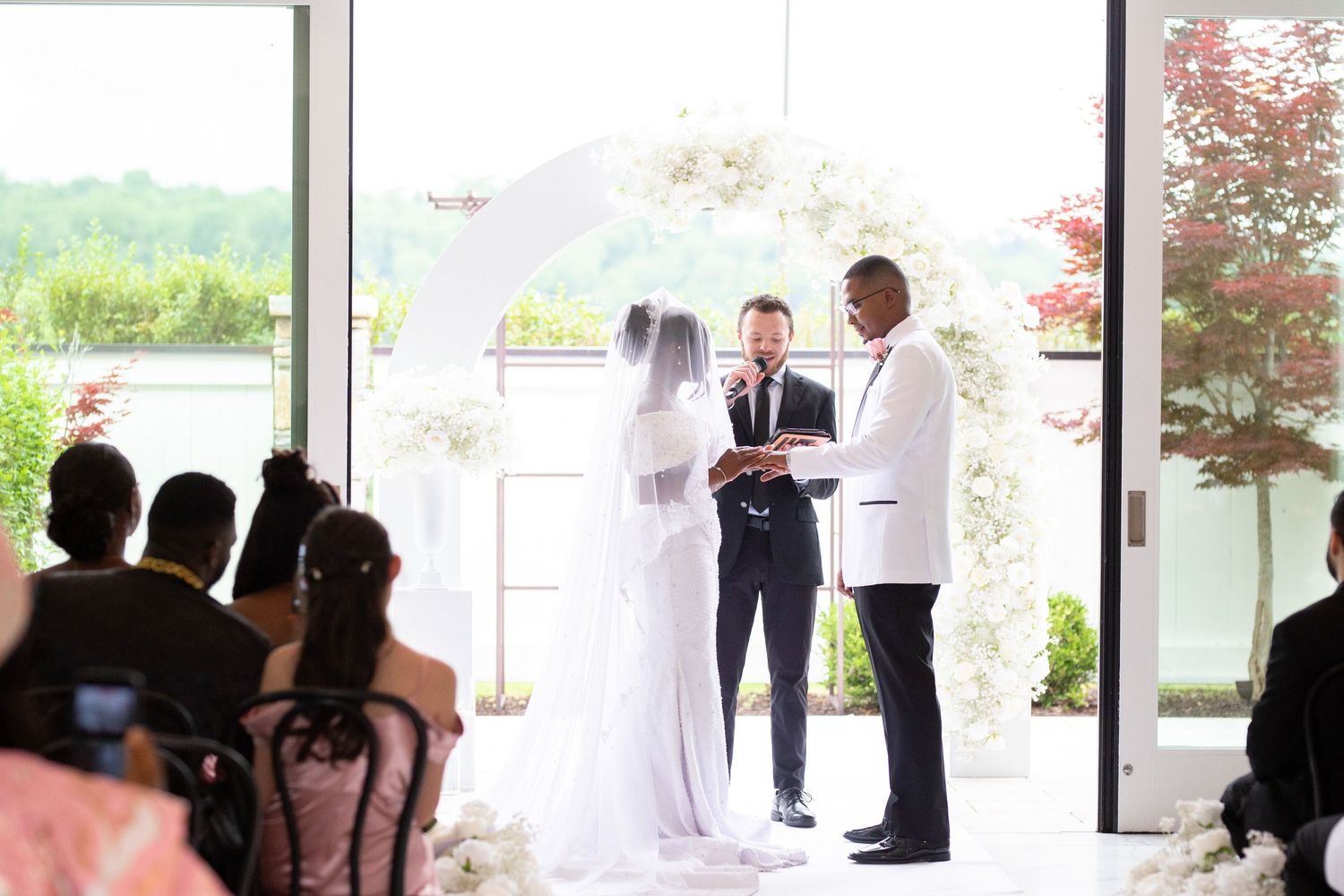 Wedding ceremony venue with white decor and large windows overlooking greenery.