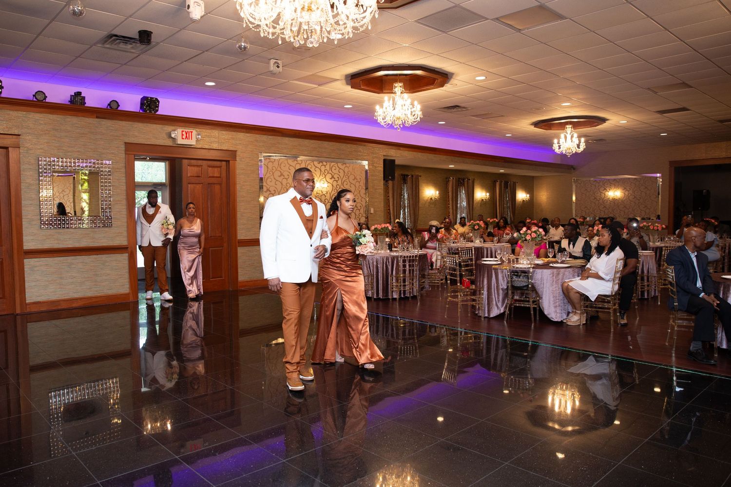 Wedding guests celebrate on the dance floor under crystal chandeliers and purple ambient lighting.