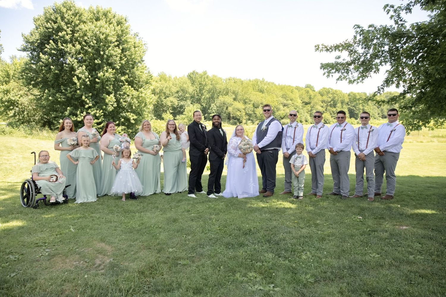 Wedding party poses outdoors on green lawn, bridesmaids in sage green gowns and groomsmen in gray suits flanking bride and groom.