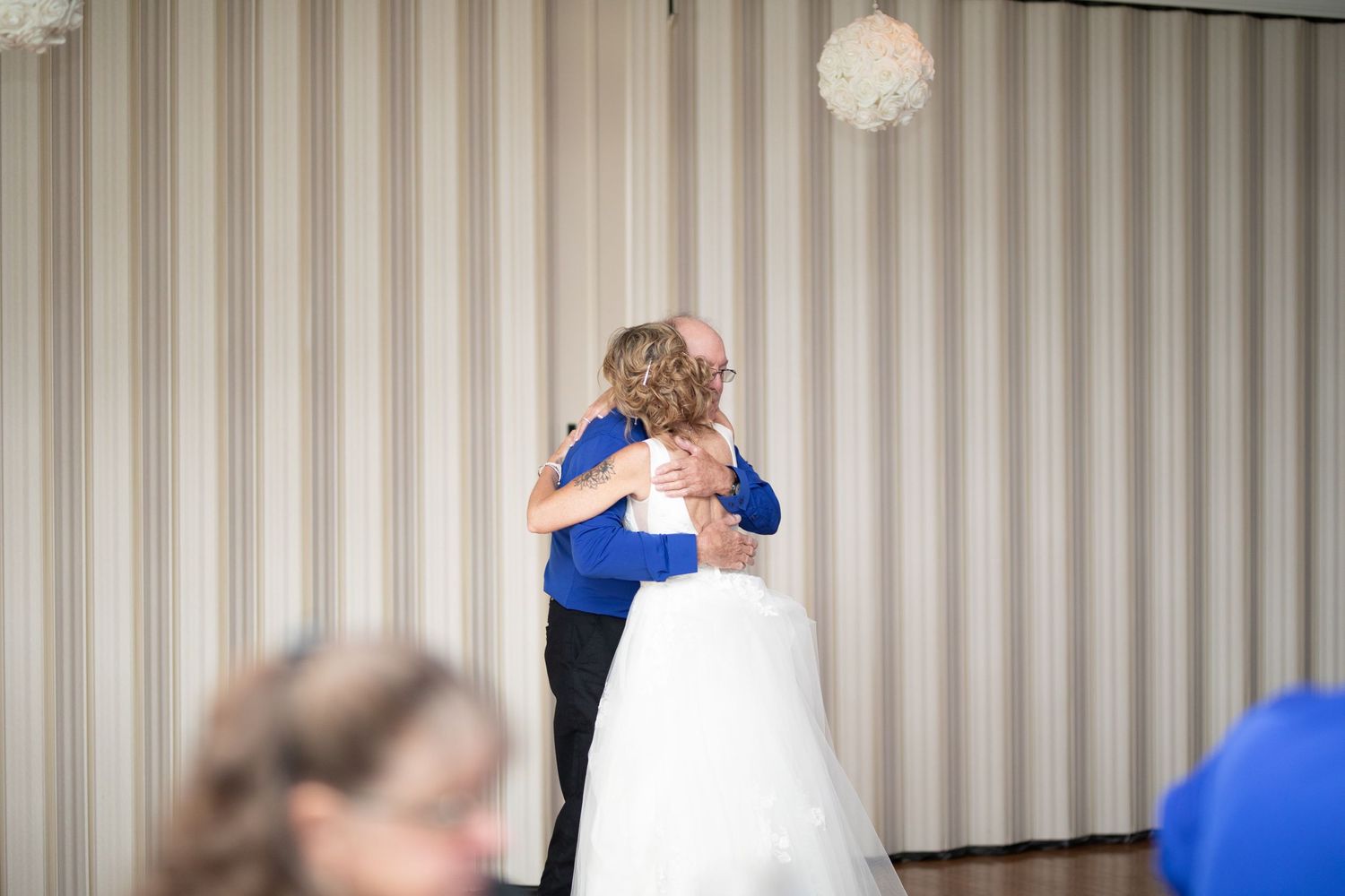Bride in white gown shares tender hug with guest in blue shirt at wedding reception.