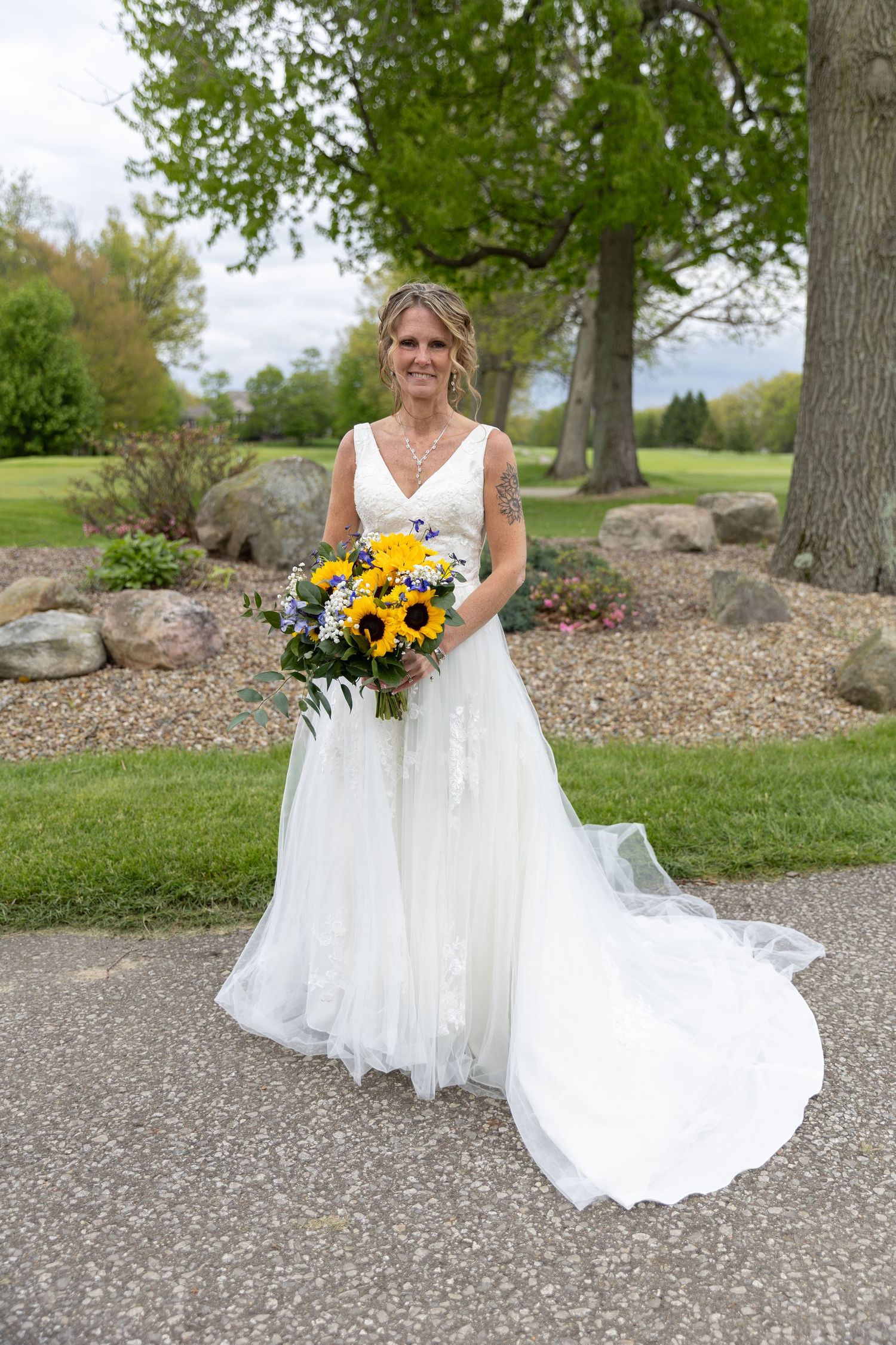 Bride in white lace gown holding sunflower bouquet, standing on garden path with trees and rocks backdrop.
