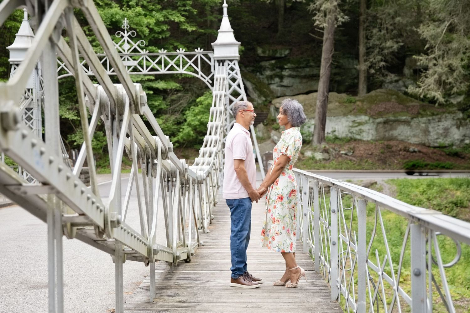 Couple holding hands on a white ornate bridge surrounded by lush green trees in a romantic outdoor setting.