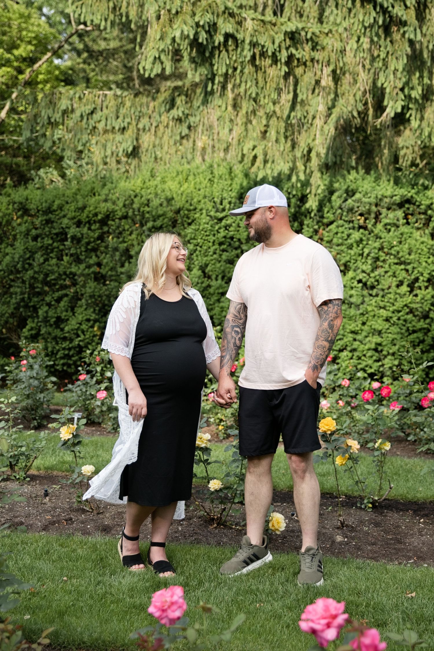Pregnant couple holding hands in a rose garden, woman in black dress and white cardigan, man in peach shirt and cap.