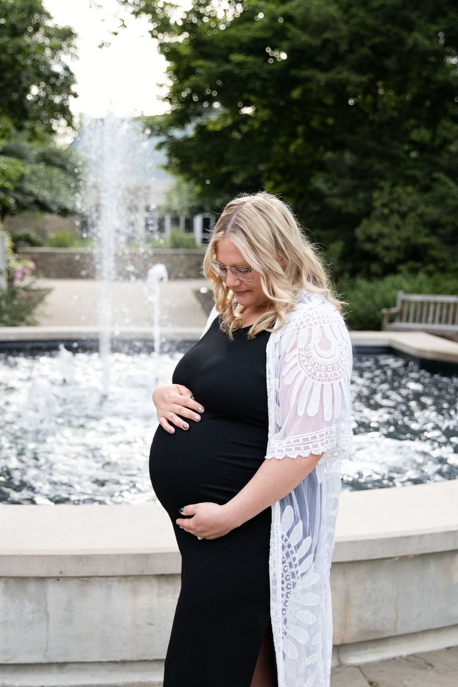 Pregnant woman in black dress and white lace cardigan cradling baby bump by a garden fountain.