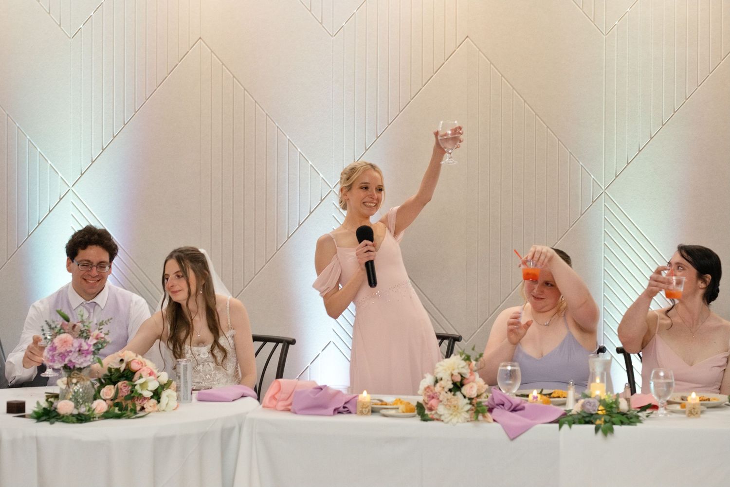 Bridesmaid gives toast raising glass at wedding head table with floral arrangements and guests.