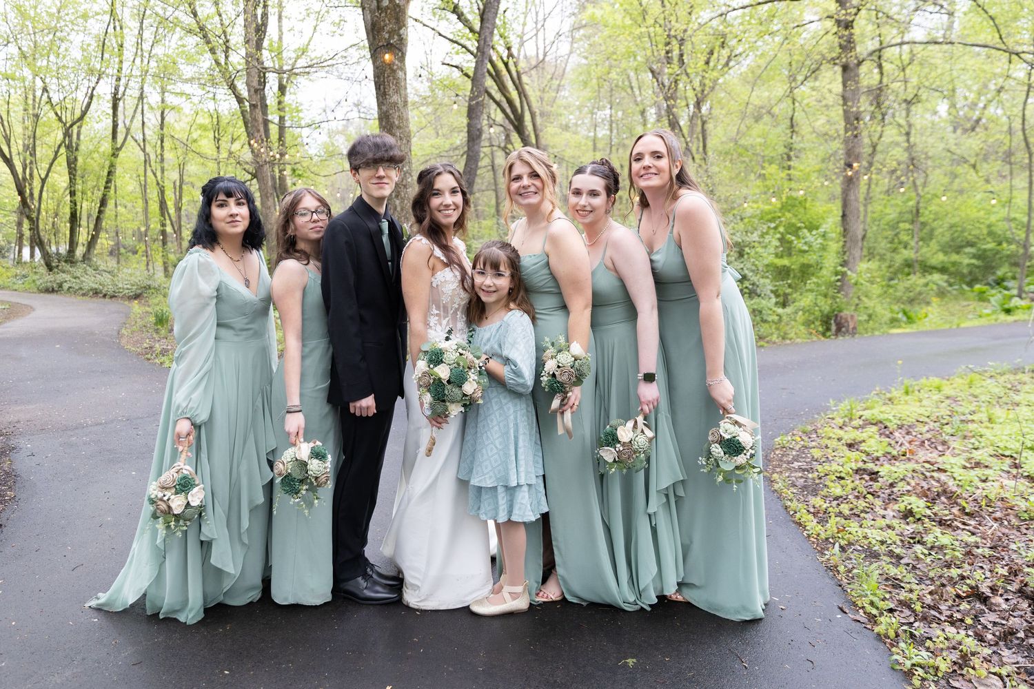 Bridal party in sage green gowns with bride in white and flower girl on a tree-lined path.
