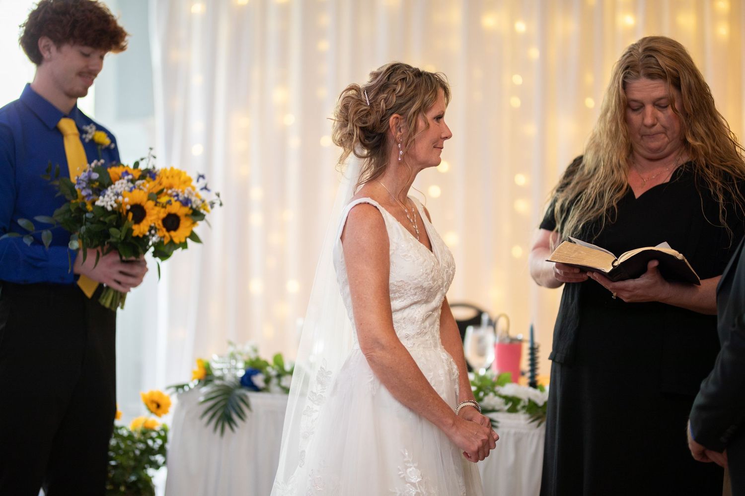 Bride in white gown stands at wedding altar with officiant reading and young groomsman holding sunflowers.