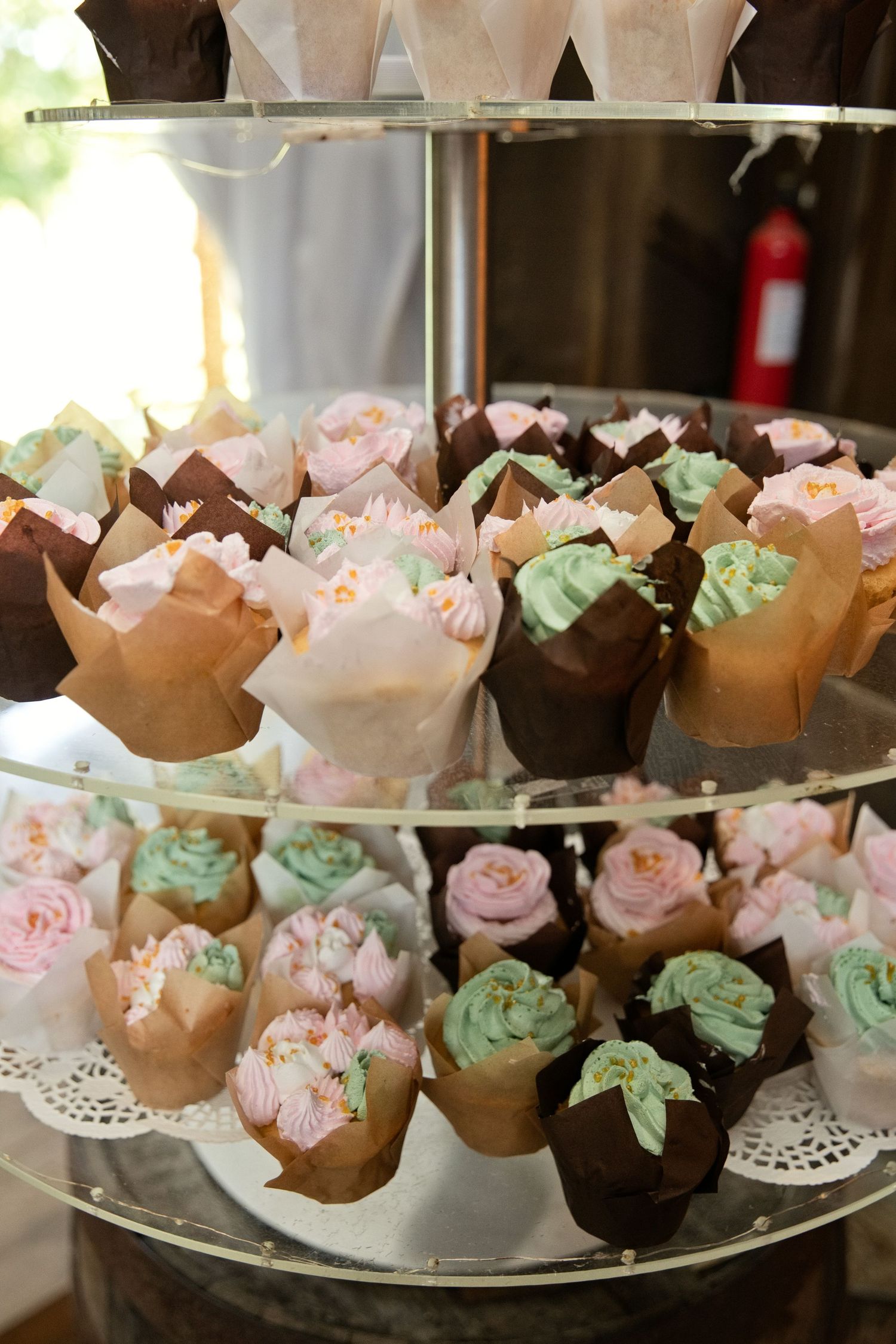 Elegant three-tier glass display stand filled with pink, mint, and white frosted cupcakes in decorative paper wrappers.