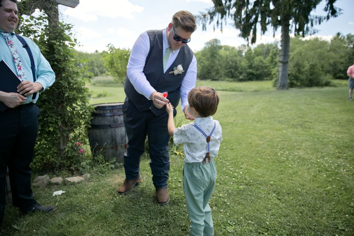 Groom in vest and sunglasses holds hands with young ring bearer in suspenders at outdoor wedding.