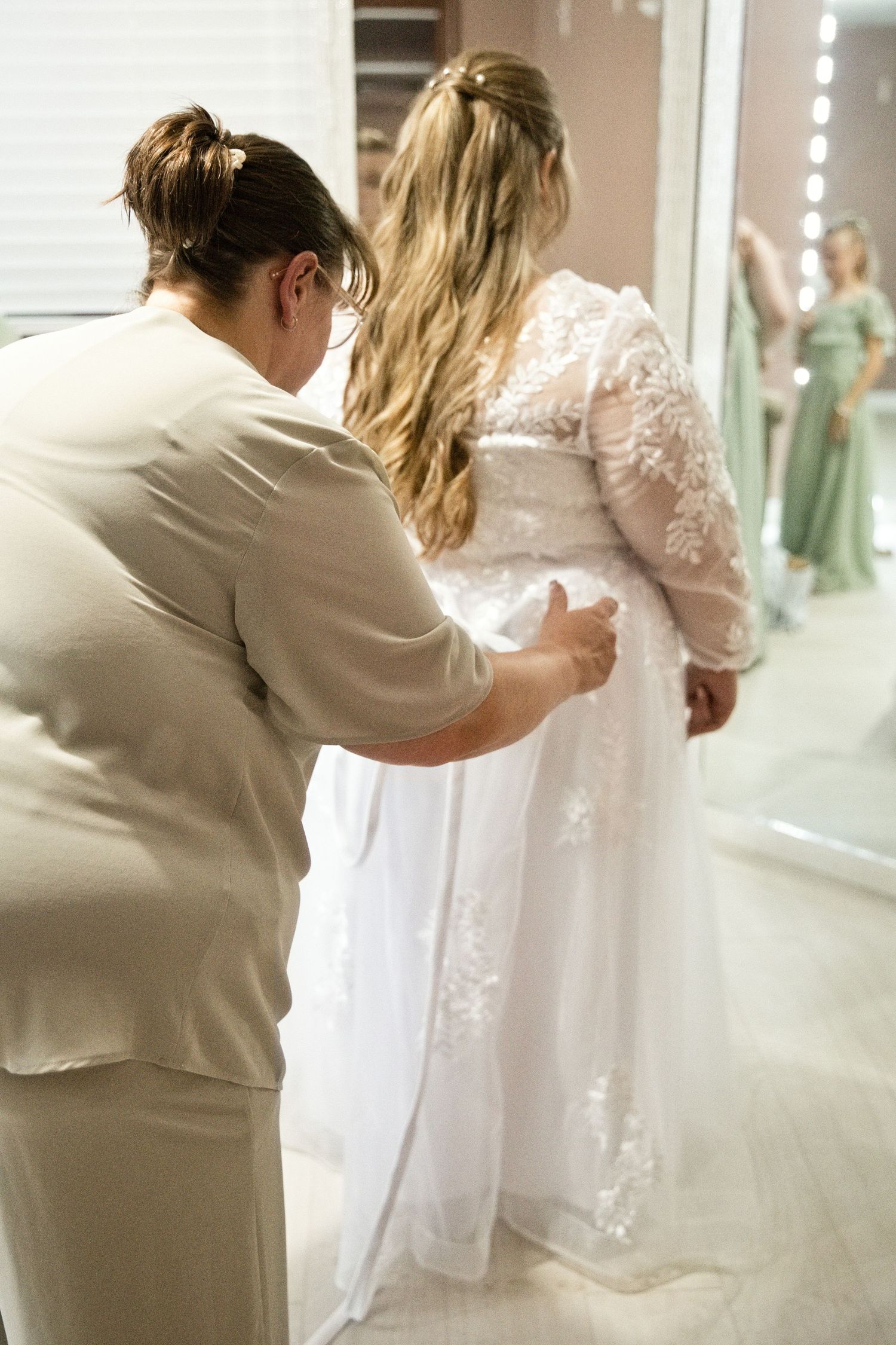 Bridal consultant helping bride into white lace wedding gown during fitting in bridal boutique.