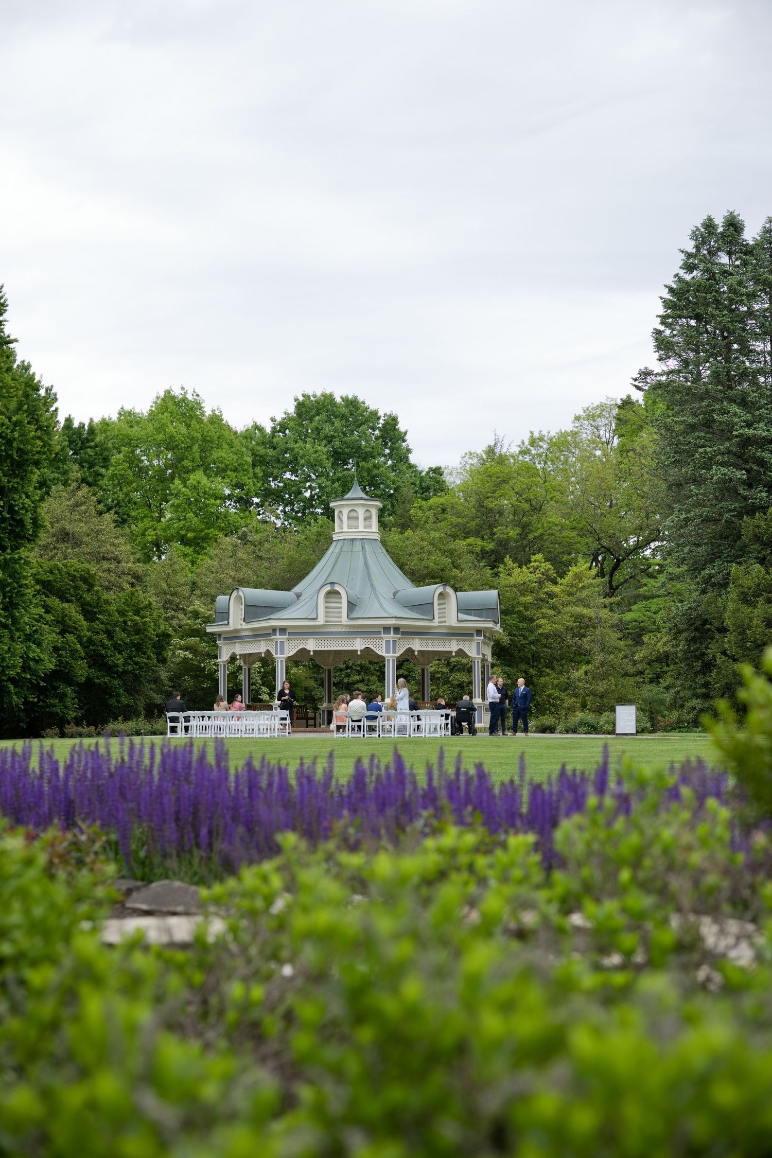 Elegant gazebo surrounded by lush green trees and purple salvia flowers in a scenic garden setting.