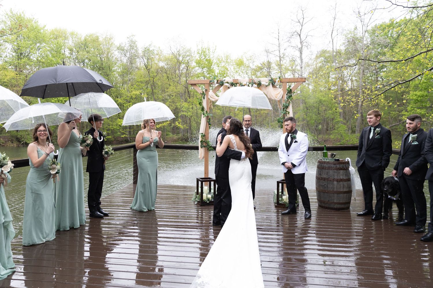 Rainy outdoor wedding ceremony on a dock with bridal party in sage green dresses holding umbrellas near a wooden arch.