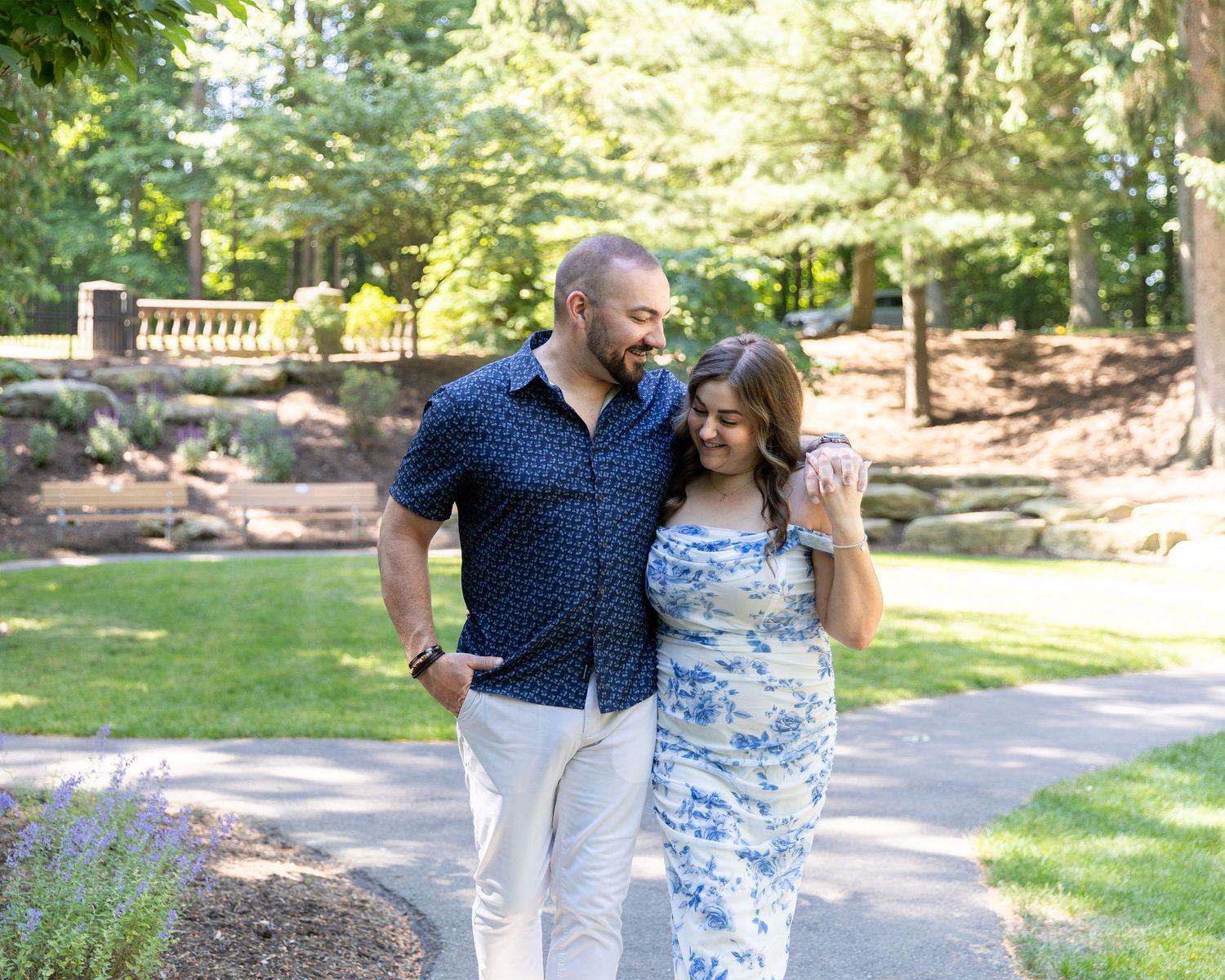 Couple walking together in a lush green park, man in navy shirt and woman in blue floral dress, smiling.