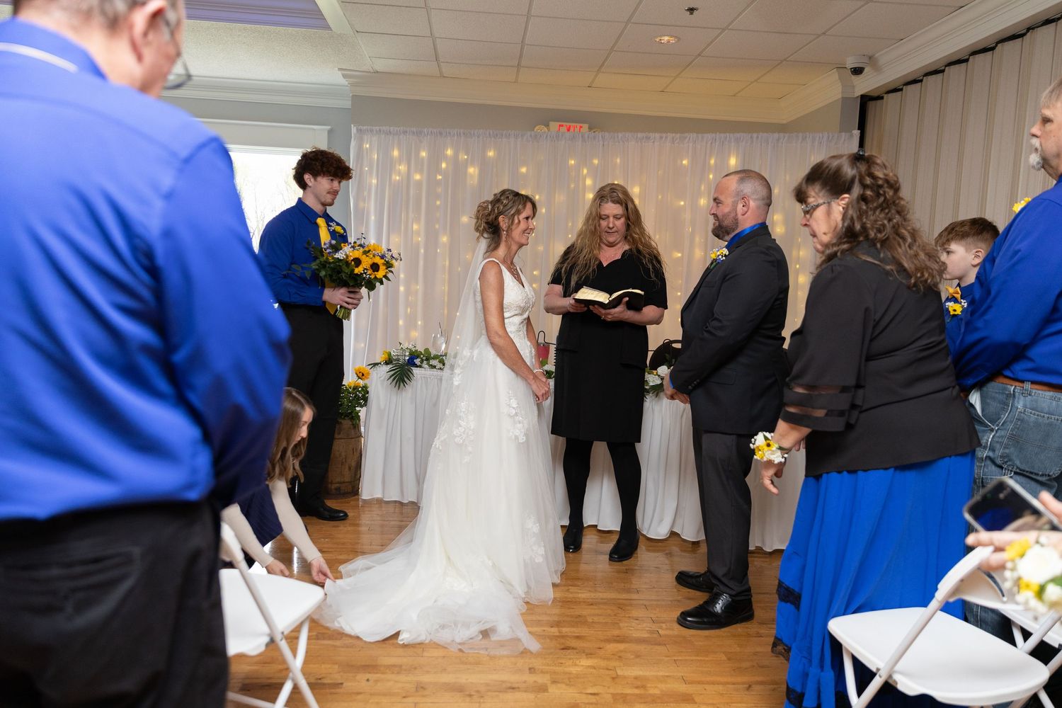 A bride and groom exchange vows at an indoor wedding ceremony with guests in blue attire and floral decorations.