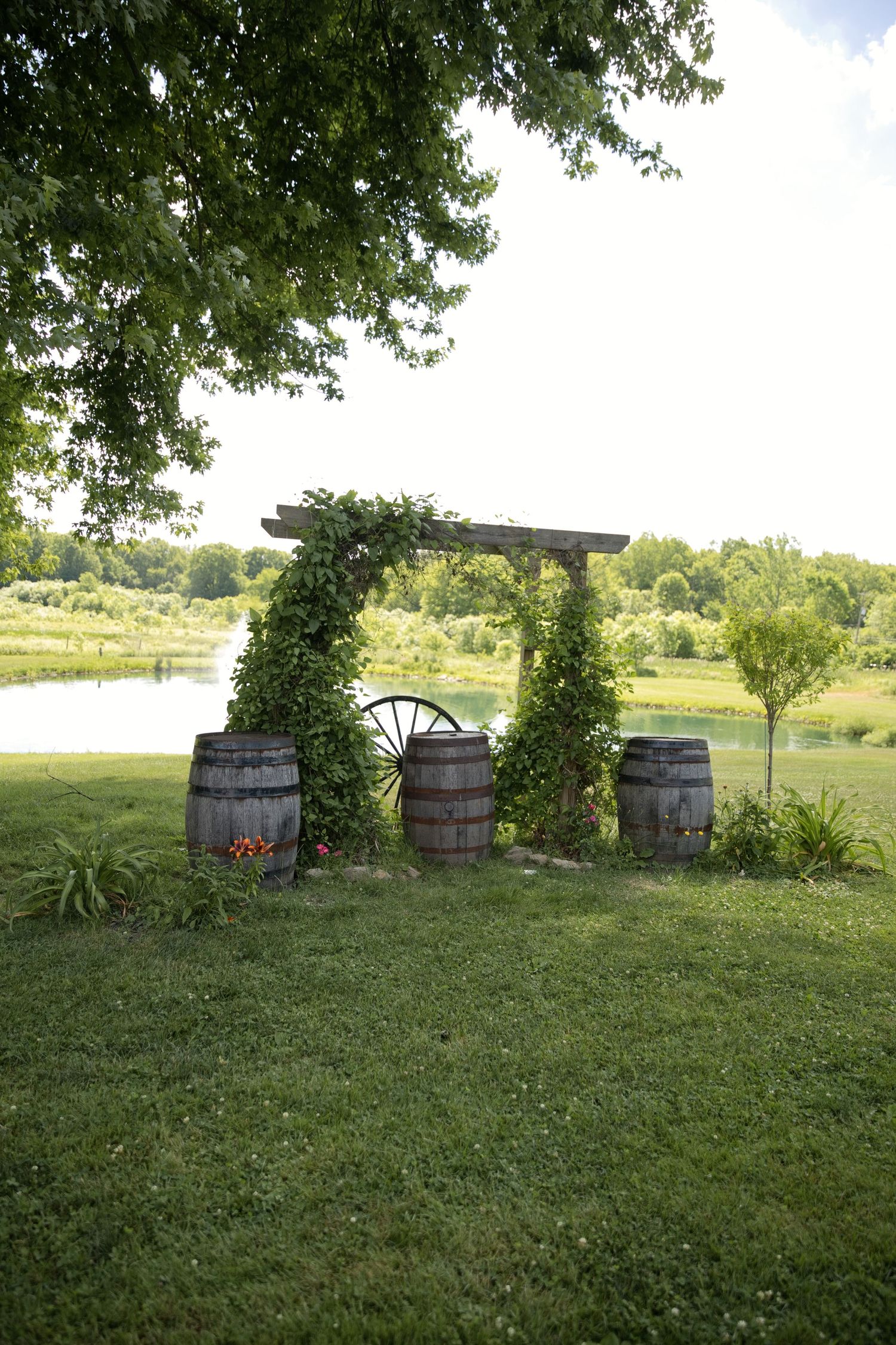Rustic wooden barrels and wagon wheel arch covered in ivy beside a serene pond, surrounded by lush green trees.