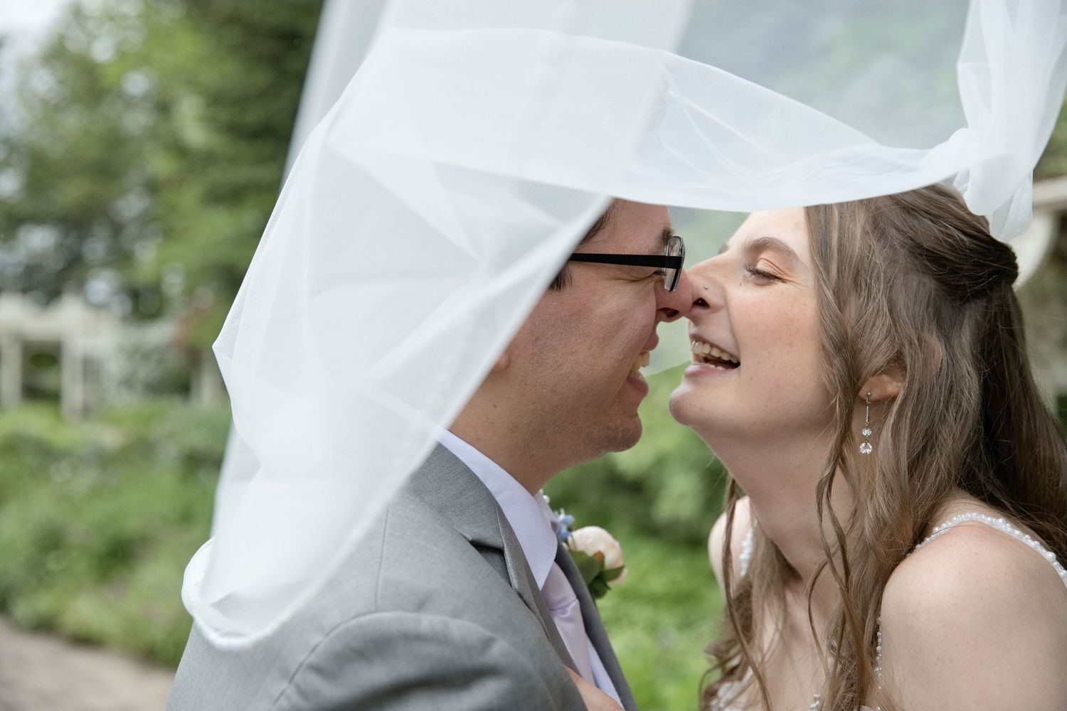 Bride and groom share a romantic nose-to-nose moment under flowing white wedding veil outdoors.