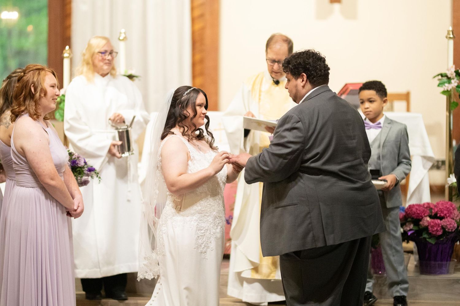 Bride and groom exchange rings during a church wedding ceremony with officiant and wedding party members present.