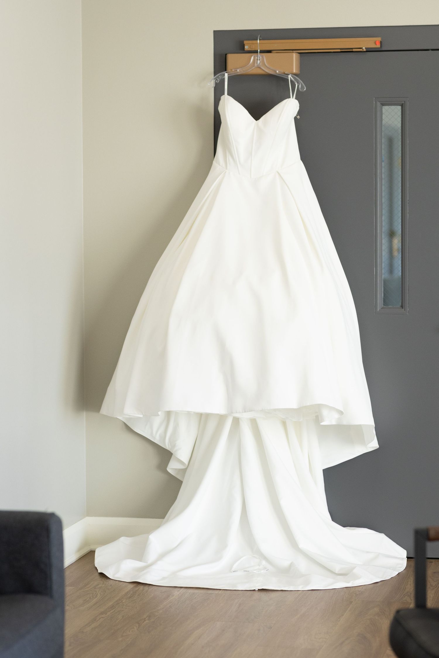 Elegant white wedding dress displayed on hanger against neutral wall.