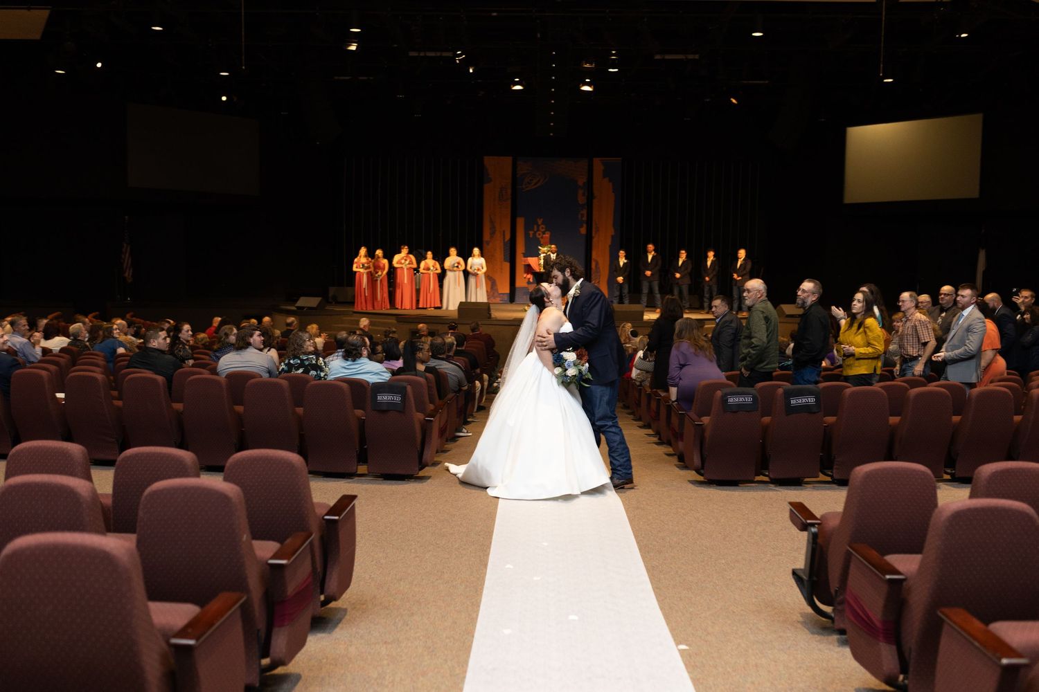 Theater wedding ceremony with white aisle runner and guests seated in burgundy chairs.