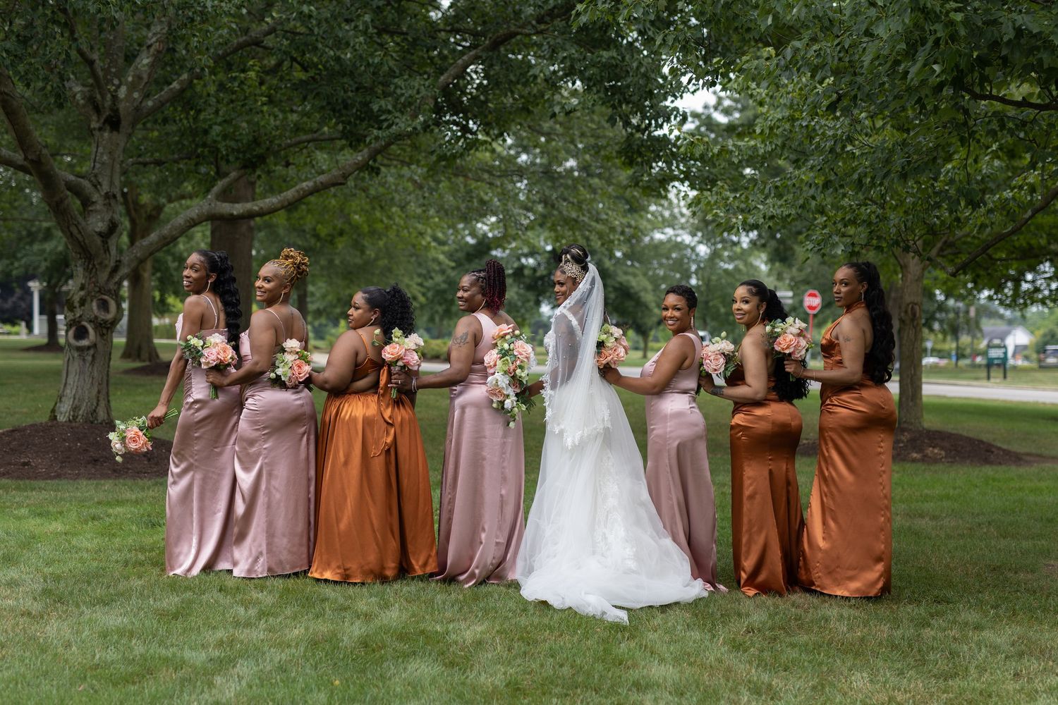 Bridal party in pink and orange dresses poses on grass under trees.