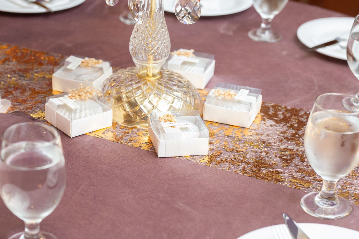 Wedding table settings with crystal glasses and favors arranged on a mauve tablecloth.