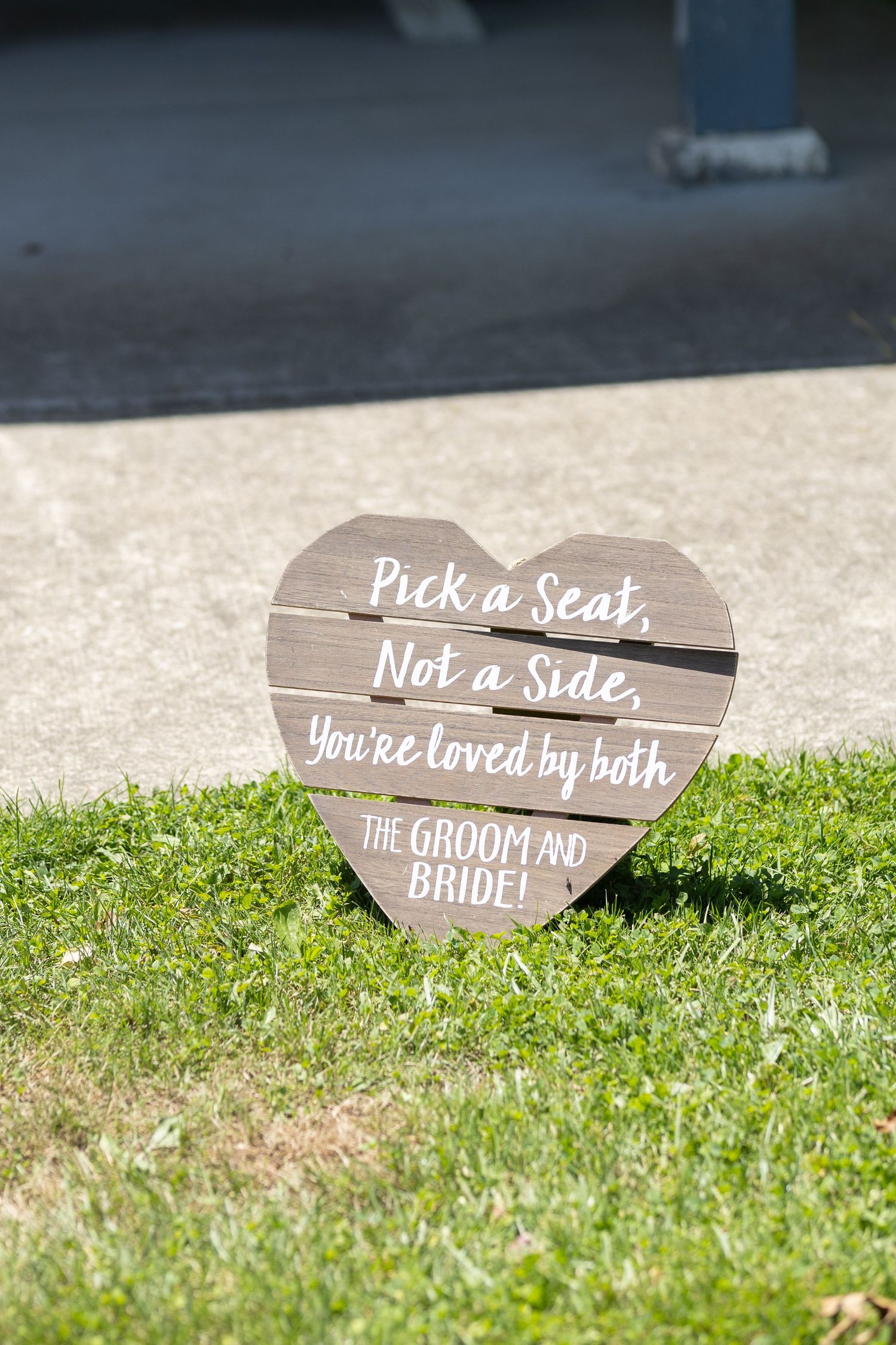 Wooden heart-shaped wedding sign on grass reads 'Pick a Seat, Not a Side'.