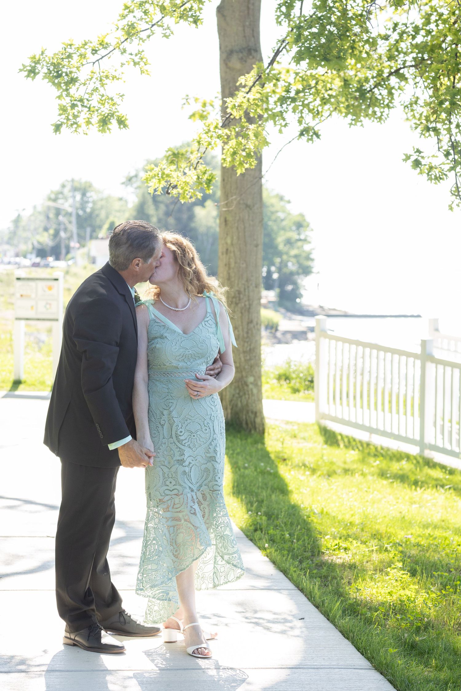 A couple shares a tender moment on a sunlit pathway bordered by green trees.