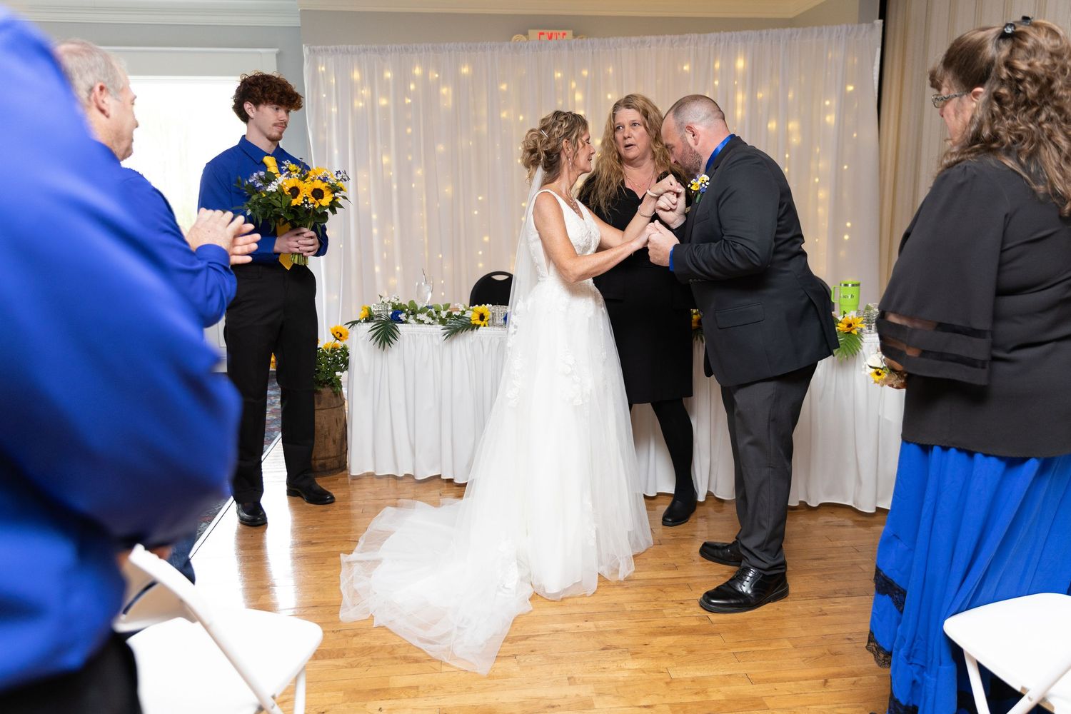 Wedding ceremony with bride in white gown, groom, and officiant exchanging rings on wooden dance floor.