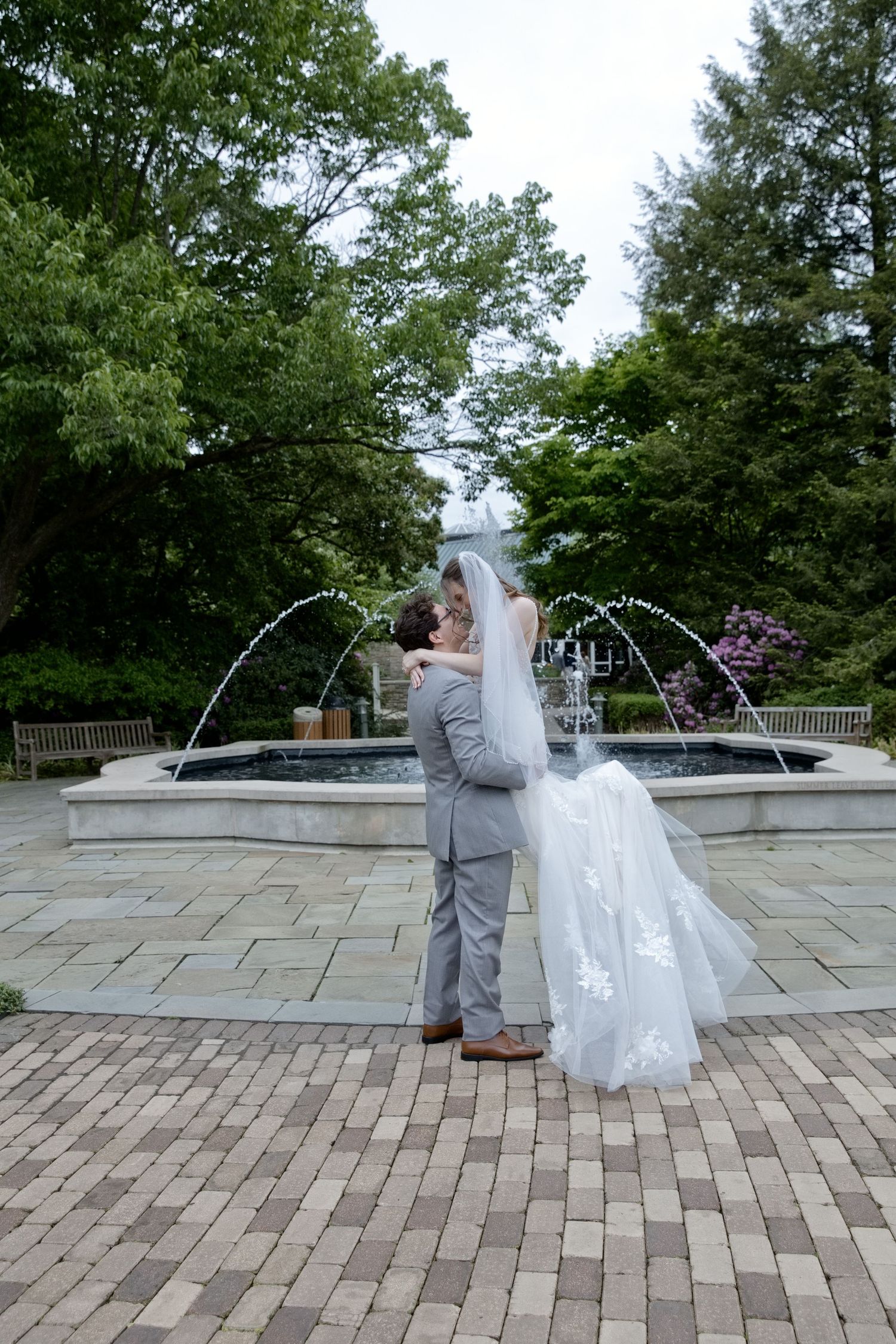 Bride and groom kissing in front of a stone fountain in a lush garden on their wedding day.
