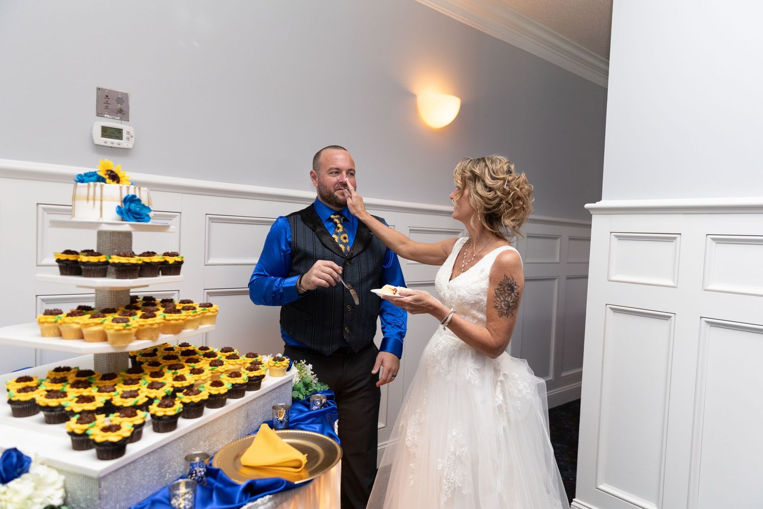 Bride feeds groom cake at wedding reception near cupcake tower with sunflowers and blue decorations.