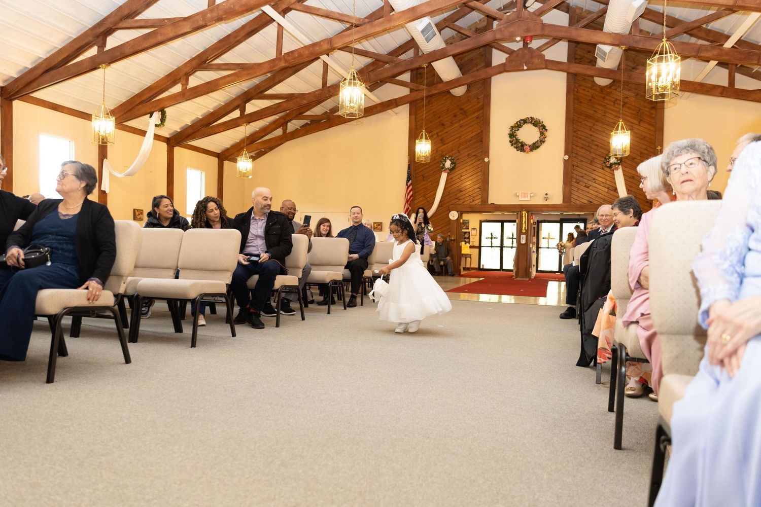 Flower girl in white dress walks down church aisle during wedding ceremony with seated guests watching.