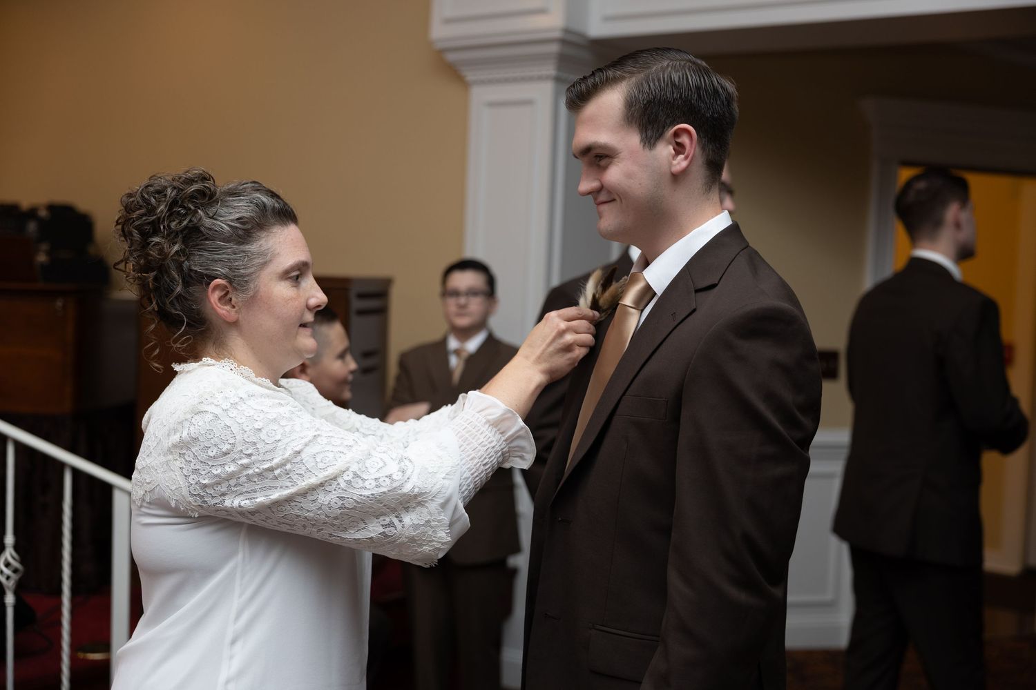 A person in white adjusts a brown tie on someone wearing a dark suit during what appears to be a formal event.