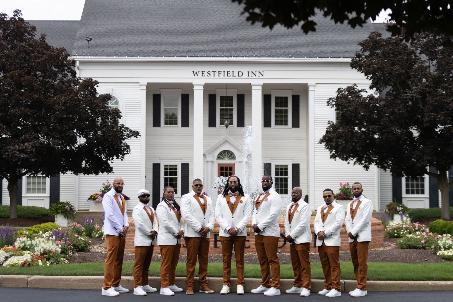 Wedding party wearing white jackets and brown pants poses in front of a white colonial style building.