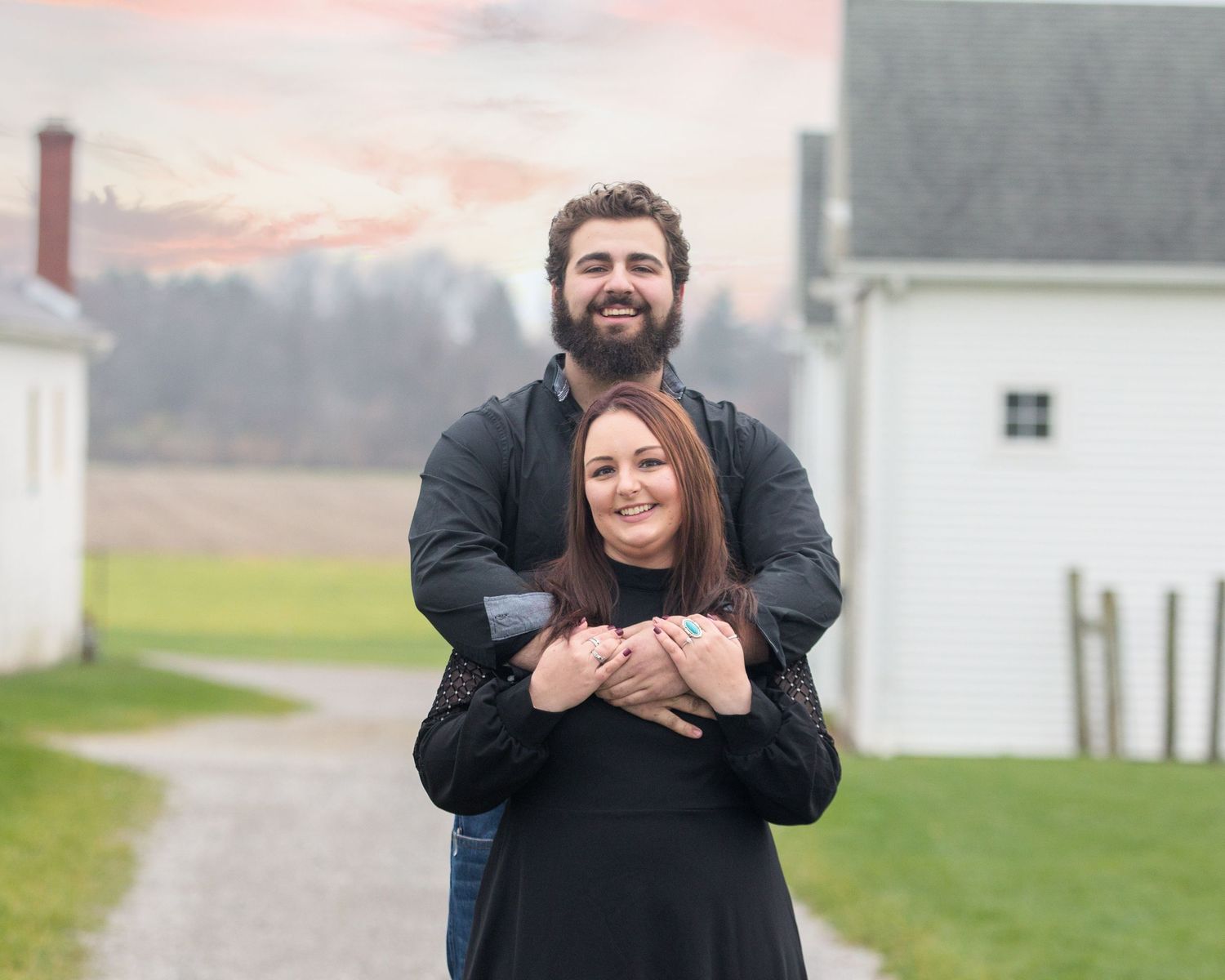 A couple embraces while standing on a gravel path with white buildings and sunset sky in background.
